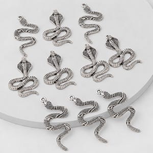 May include: A collection of ten silver-toned metal snake charms. The charms feature two distinct designs: some depict coiled cobras with flared hoods, while others show slender, winding snakes in a serpentine pose. Each charm has a small loop at the top.