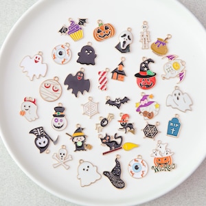 Op de afbeelding: Een verzameling diverse Halloween-bedels van metaal met emaille details, waaronder ontwerpen zoals geesten, pompoenen, skeletten, oogballen, zwarte katten, heksen, vleermuizen, spinnenwebben, grafstenen en snoepjes in diverse kleuren.