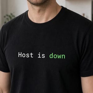 Puede incluir: Una camiseta negra de manga corta y cuello redondo con el texto "Host is down" impreso en el pecho. Las palabras "Host" e "is" son de color blanco, mientras que la palabra "down" es de un color verde brillante. La prenda tiene un corte estándar.