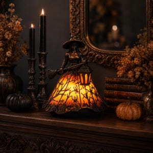 Op de afbeelding: Een decoratieve lamp in de vorm van een heks met een puntmuts die een lantaarn vasthoudt. De voet van de lamp heeft een doorschijnende oranje kap versierd met zwarte silhouetten van kale boomtakken en spinnen, die een warme gloed uitstraalt.