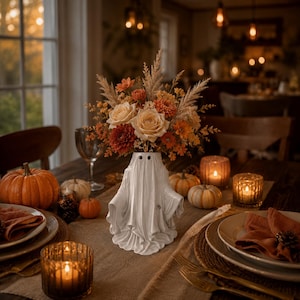 May include: A white ceramic ghost-shaped vase filled with a bouquet of dried flowers in shades of orange, cream, and brown. The ghost figure features two black circular eyes and a draped, flowing silhouette, serving as a centrepiece on a table.