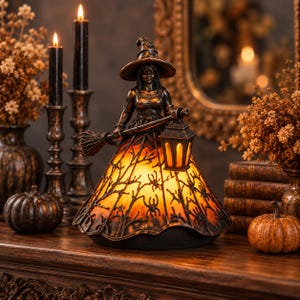 May include: A decorative witch figurine lamp featuring a dark bronze-toned figure wearing a pointed hat. The witch holds a broom and a lantern, while the skirt is illuminated from within, displaying a black silhouette pattern of spiders and branches.