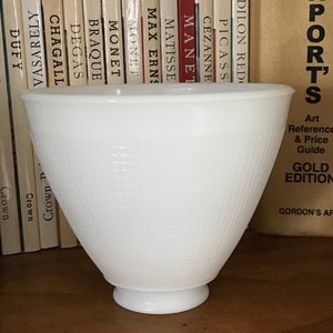 May include: A white glass bowl with a textured, grid-like pattern. The bowl has a flared design and a pedestal base. The background features books on a shelf and a gold-coloured book.