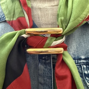 May include: A silk scarf featuring a pattern of green, red, and black stripes, secured by two polished gold-tone metal bars. The scarf is draped over a blue denim shirt, creating a layered look with the vibrant fabric colors and metallic accents.