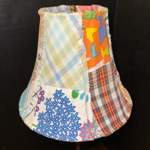 Op de afbeelding: Een patchwork lampenkap gemaakt van verschillende stoffen in verschillende kleuren en patronen, waaronder ruit, bloemen en geometrische patronen.