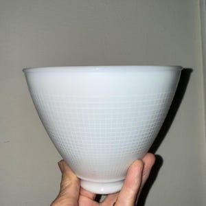 May include: A white milk glass lamp shade with a conical shape and a subtle grid pattern etched across the exterior surface. The shade features a narrow circular base opening and a wide, flared top opening with a smooth, rounded rim edge.