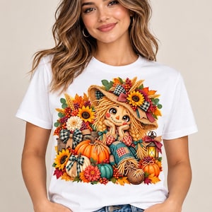 May include: A white t-shirt featuring a colorful graphic print of a scarecrow wearing a straw hat, surrounded by autumn elements including orange and white pumpkins, sunflowers, red berries, green leaves, and a glowing lantern.