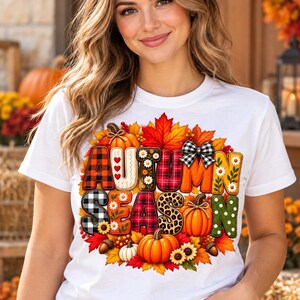 May include: A white short-sleeved t-shirt featuring a colorful graphic print that reads AUTUMN SEASON. The letters are decorated with various patterns including plaid, leopard print, and floral designs, surrounded by pumpkins and autumn leaves.
