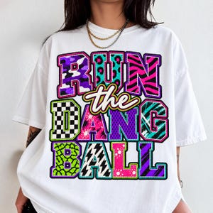 Könnte beinhalten: Ein weißes T-Shirt mit dem Schriftzug RUN THE DANG BALL in großen, blockartigen Buchstaben. Jeder Buchstabe zeigt ein anderes lebhaftes Muster, darunter lila Camouflage, türkisfarbener Leopardenprint, pinke Zebrastreifen und schwarz-weiße Karos.
