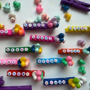May include: A collection of colorful wooden clothespins, each featuring the word STUNT spelled out in white letter beads. Several small, multi-colored miniature rubber ducks are attached to the ends of the clips, with additional loose ducks scattered around the arrangement.