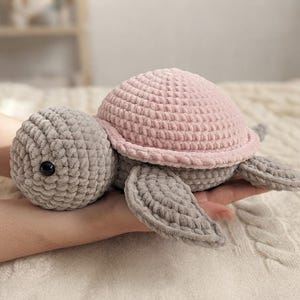Könnte beinhalten: Ein gehäkeltes Schildkröten-Stofftier mit einem weichen, strukturierten grauen Kopf und Flossen sowie einem abgerundeten, blassrosa Panzer. Die Schildkröte hat kleine, runde, schwarze Kunststoffaugen, die in den grauen Garnkopf eingearbeitet sind.