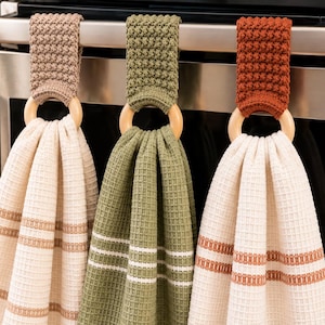 May include: Three hanging kitchen towels with waffle-weave fabric, each attached to a light-colored wooden ring. The tops feature textured crochet toppers in beige, olive green, and rust orange, matching the striped accents on the towels.