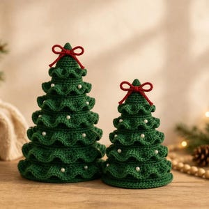 Könnte beinhalten: Zwei kegelförmige Weihnachtsbäume aus dunkelgrünem Häkelgarn. Jeder Baum verfügt über gestufte, gerüschte Lagen, kleine weiße perlenartige Kügelchen auf dem Stoff und eine kleine rote Garnschleife an der obersten Spitze.