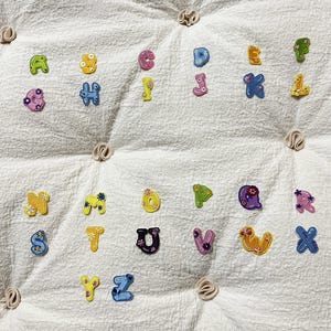 May include: A collection of twenty-six individual alphabet letter patches, each featuring a unique colour and floral pattern. The embroidered letters range from A to Z, showcasing various shades of blue, yellow, pink, green, and purple.