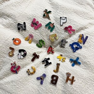 Puede incluir: Una colección de veintiséis parches bordados con letras del alfabeto, cada uno con un diseño único de animal u objeto. Los coloridos parches incluyen letras como la K con un canguro, la M con un mono, la R con una rosa y la U con un unicornio, mostrando formas diversas y colores vibrantes.