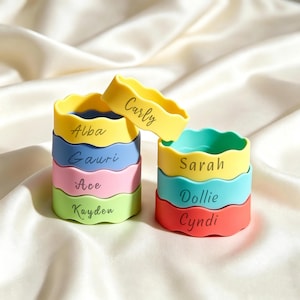 Op de afbeelding: Een verzameling van acht siliconen armbanden met golvende randen, gestapeld in twee torens. De armbanden zijn geel, blauw, roze, lichtgroen, turquoise en koraalkleurig, elk voorzien van een in zwarte schrijfletters gegraveerde naam: Carly, Alba, Gauri, Ace, Kayden, Sarah, Dollie en Cyndi.