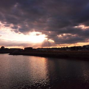 Könnte beinhalten: Ein Sonnenuntergang über einem Gewässer mit dunklen, schweren Wolken, die die Sonne teilweise verdecken und helle Lichtstrahlen erzeugen, die sich auf der gekräuselten Wasseroberfläche spiegeln. Der Horizont zeigt die Silhouette von Gebäuden.