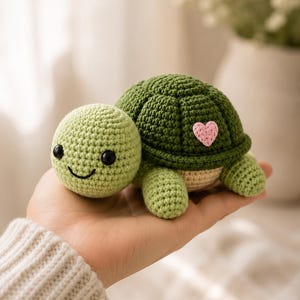 May include: A small, handmade crochet turtle toy featuring a light green head with black bead eyes and a stitched smile. The turtle has a dark green shell adorned with a small, light pink crocheted heart patch on the side and a beige underside.