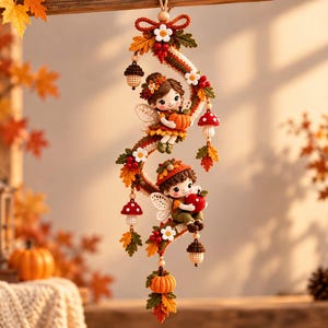 Op de afbeelding: Een hangende gehaakte decoratie met twee gevleugelde feeënfiguren, waarvan er één een pompoen vasthoudt en de andere een appel. De structuur is versierd met herfstachtige elementen zoals oranje en gele bladeren, rode bessen, witte bloemen, bruine eikels en rood-witte paddenstoelen.