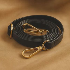 May include: A black bag strap coiled in a circular shape, featuring polished gold-tone metal lobster claw clasps at both ends. The strap is constructed with smooth, dark material and includes matching gold-tone metal hardware rings for attachment.