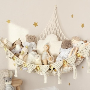 May include: A cream-colored macrame toy hammock featuring a triangular net design with decorative tassels and small pennant flags. The hammock is filled with various plush toys, including teddy bears, dolls, and a white cloud-shaped pillow.