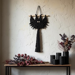 May include: A black macrame wall hanging shaped like a bat, suspended from a light-colored wooden dowel. The design features two small, round, light-colored wooden beads as eyes and a long, thick fringe tassel hanging from the bottom center.