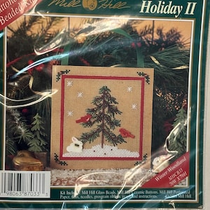 May include: A Mill Hill Holiday II Winter Woodland buttoned and beaded craft kit in a plastic package. The kit features a square perforated paper design of a green pine tree, red birds, and a white rabbit on a tan background with snow.