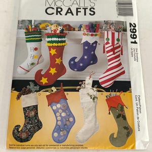 Może przedstawiać: Koperta z wykrojem krawieckim McCall's Crafts 2991 z ilustracjami ośmiu różnych skarpet świątecznych. Wzory obejmują różne kolory, wzory i elementy dekoracyjne, takie jak pompony, guziki i tekstury tkanin do dekoracji świątecznych.