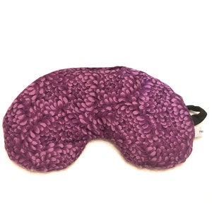 May include: Purple crescent-shaped eye pillow with a floral pattern. The pillow has a black drawstring closure.