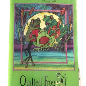 Könnte beinhalten: Eine grüne Verpackung mit einem Wandquilt-Muster mit dem Titel "1012-Frog Choir". Die Illustration zeigt drei grüne Frösche, von denen einer ein Saiteninstrument spielt. Die Verpackung enthält auch den Text "Quilted Frog" und "Patterns for Quilters Copyright © 2004."