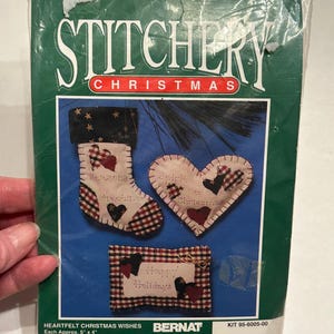 May include: A craft kit for making three felt Christmas ornaments. The kit includes a red and white checked stocking, a heart-shaped ornament with the text "Christmas" and a rectangular ornament with the text "Happy Holidays". The kit is from Bernat Crafts and is labelled "Stitchery Christmas".