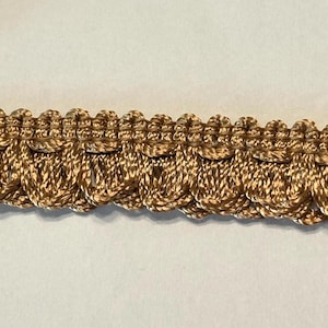 May include: Close-up of a decorative gold trim with a looped design. The trim is made of a woven material and has a textured appearance. The trim is suitable for various craft and sewing projects.
