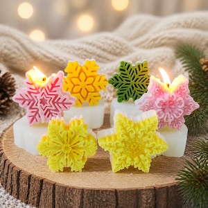 Può includere: Una collezione di sei candele in cera a forma di fiocco di neve nei colori rosa, giallo e verde, ciascuna poggiata su una base bianca. Due candele sono accese con una fiamma calda. Sono disposte su una fetta di legno circolare con corteccia.