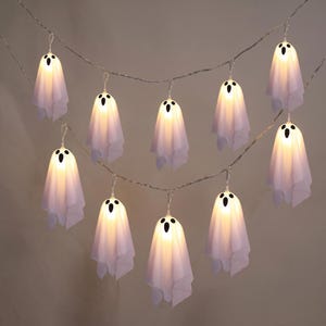 May include: A string of ten white ghost-shaped LED lights hanging in two rows. Each ghost is made of a thin, draped fabric with two black oval eyes and a black oval mouth printed on the front, illuminated from within by a warm glowing light.