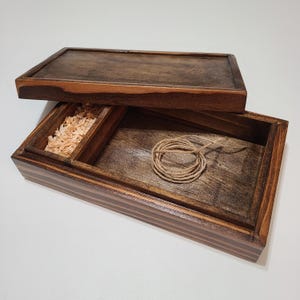 May include: A wooden box with a lid, featuring a dark brown finish. The box has a compartment with wood shavings and another with a coiled twine. The lid is slightly ajar, revealing the interior.