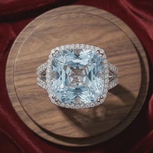 Op de afbeelding: Een zilverkleurige ring met een grote, vierkante, lichtblauwe edelsteen in kussenslijpsel in het midden. De steen is omringd door een halo van kleine, heldere, rondgeslepen stenen, met extra kleine steentjes langs de gespleten ringband.