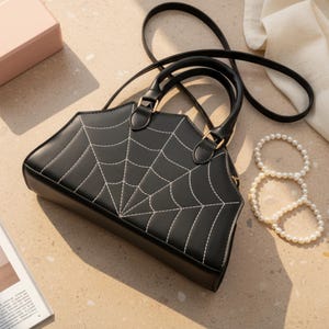 Könnte beinhalten: Eine schwarze Handtasche in strukturierter Sargform mit weißem, aufgesticktem Spinnennetz-Design auf der Vorderseite. Die Tasche verfügt über zwei kurze Tragegriffe und einen langen, schmalen Schulterriemen mit goldfarbenen Beschlägen.