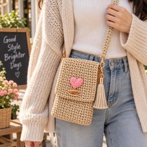 May include: A beige crochet crossbody bag featuring a small pink heart appliqué on the front flap, a gold-toned metal tag labeled handmade, and a matching crochet strap with a long beige tassel attached by a gold-toned metal swivel clasp.