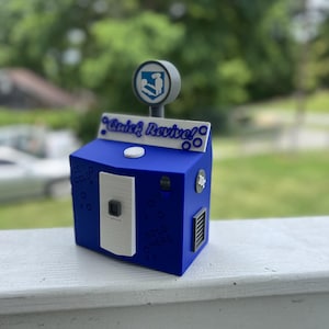 May include: A blue vending machine replica featuring the text Quick Revive, a circular sign with a white icon, a white central panel, and small circular details. The machine includes text reading ICE COLD and SOLD HERE, with a 10c coin slot.