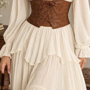 May include: A long-sleeved cream-colored dress with a square neckline and a tiered skirt, featuring a brown corset-style waist belt with a lace-up front closure and a subtle embossed floral pattern, paired with a small brown handbag.
