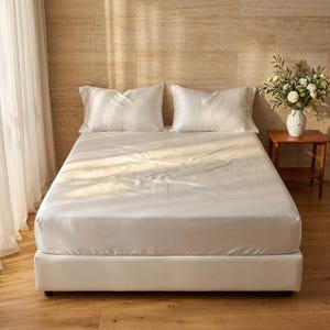 Può includere: Un letto con un lenzuolo con angoli bianco liscio e due federe bianche abbinate. Il letto è posizionato in una stanza con pareti in pietra chiara e pavimento in legno, con un vaso bianco contenente fiori bianchi su un tavolino.