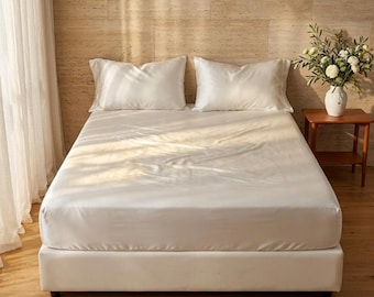 Könnte beinhalten: Ein Bett mit einem glatten, einfarbig weißen Spannbettlaken und zwei passenden weißen Kopfkissenbezügen. Das Bett steht vor einer hellen Steinwand, daneben ein weißer Vasenbehälter mit grünem Blattwerk und weißen Blumen auf einem Holztisch.