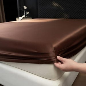 May include: A solid dark brown fitted sheet with a smooth, satin-like finish covering a mattress. The fabric has a subtle sheen and is pulled taut over the corner of the bed, showing the deep chocolate color and the texture of the material.