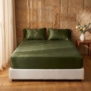 May include: A bed featuring a smooth, olive green fitted sheet and two matching olive green pillowcases. The bed is set against a textured stone wall, with a white base frame and a vase of flowers on a small wooden side table nearby.