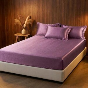 May include: A bed featuring a smooth, solid purple sheet set with a fitted sheet and three matching rectangular pillows. The bed is positioned against a dark wood paneled wall, with a small wooden side table holding a vase of dried grass.