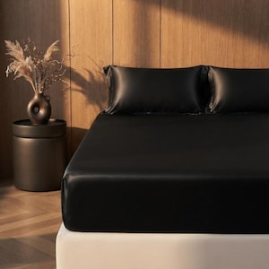 May include: A set of smooth, solid black bed sheets and two matching pillowcases covering a mattress. The fabric has a subtle sheen, creating a uniform dark appearance across the bedding surface, providing a clean and consistent look for the bed.