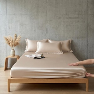 May include: A champagne-colored satin bed sheet set featuring a fitted sheet, a flat sheet, and two matching pillowcases with decorative flange edges. The smooth, lustrous fabric covers a wooden bed frame, creating a sleek and uniform appearance.