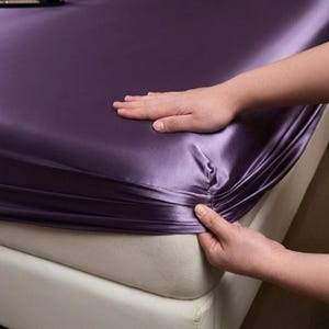 May include: A deep purple fitted sheet with a smooth, lustrous finish. The fabric is pulled taut over the corner of a mattress, showcasing the elasticized edge and the reflective, satin-like texture of the material in a rich, dark violet hue.