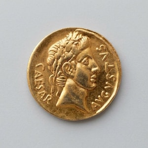 May include: A circular gold coin featuring a profile relief of a person wearing a laurel wreath. The text CAESAR is inscribed on the left side and the text AVGVSTVS is inscribed on the right side of the coin's face.