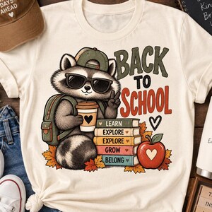 Puede incluir: Una camiseta color crema con un gráfico de un mapache con gorra verde y gafas de sol, sosteniendo una taza de café y haciendo el signo de la paz. El texto dice BACK TO SCHOOL, LEARN, EXPLORE, GROW, BELONG, junto a libros y una manzana roja.